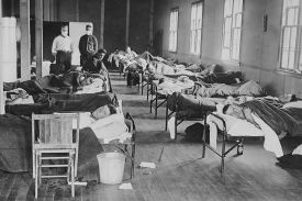 A black-and-white historical photograph of a crowded hospital ward. Rows of patients lie in narrow metal cots covered with blankets, while two individuals wearing white face masks stand at the far end of the room.