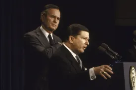 A man in a dark suit speaks at a podium with two microphones, his right hand resting on the side of the lectern which features a circular seal. Standing slightly behind him and to the left, another man in a grey suit and tie watches with a serious expression. The background consists of dark blue curtains and dim lighting.