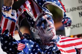 A close-up shot shows a man dressed like a medieval court jester, with a shirt and dangly hat in an American flag pattern. The man's face is pointed white with a red line down the middle and blue smudges around his eyes.