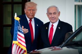 Trump and Netanyahu stand shoulder to shoulder as they pose for media. Netanyahu smiles slightly; Trump has a serious expression and gestures with one hand. The hood of a vehicle with a small American flag displayed on it is visible in the foreground