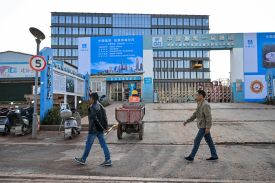 Two men walk on a sidewalk as they pass by a mostly-completed midrise building with shiny windows. Large posters and signage with writing in Chinese stand in front of the complex, describing the project. A man heading toward the building pulls a wheelbarrow behind him.