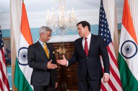 Two men shake hands, with the Indian and American flags beside them.