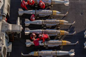 About half a dozen U.S. Navy sailors are seen from overhead as they work on what appear to be torpedos or missiles of some sort. The sailors wear bright red uniforms and helmets.