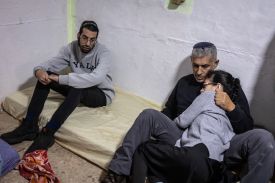 Two men sit on a mattress on the floor in a corner of a bunker. The older man cradles a young woman who rests her head on his shoulder, whereas a younger man at his side simply stares.