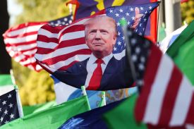 An array of flags are seen billowing in the wind, including Iranian flags, American flags, and a large American flag embellished with a cut-out photo of Donald Trump giving a thumbs up.