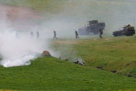 Soldiers in full combat gear and helmets perform a tactical exercise on a green, hilly field. Thick white smoke billows from a canister in the foreground, partially obscuring the view. Some soldiers run across the open grass toward military vehicles, while others are positioned in a shallow trench, aiming their rifles.