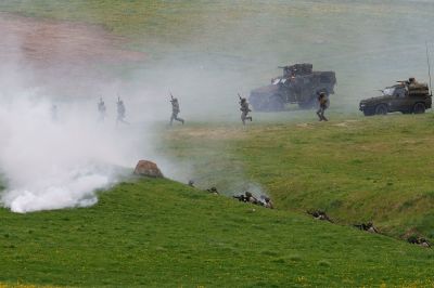 Soldiers in full combat gear and helmets perform a tactical exercise on a green, hilly field. Thick white smoke billows from a canister in the foreground, partially obscuring the view. Some soldiers run across the open grass toward military vehicles, while others are positioned in a shallow trench, aiming their rifles.