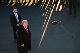 Trump, in a black overcoat and red tie, is seen from above as he speaks into a boom mic, while Mullin stands behind him with hands clasped. At the top of the image one can see the feet of spectators standing in a line, letting in beams of sunlight through the gaps between their legs.