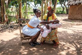 A midwife in a white uniform with an orange belt sits outdoors, leaning forward to examine a newborn baby held by a woman in a yellow shirt and colorful patterned skirt. They are seated in a sunlit, dirt clearing surrounded by trees and a traditional thatched-roof building in the background.