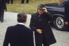 Mikhail Gorbachev, wearing a dark overcoat and patterned scarf, smiles as he tips his gray fedora toward Ronald Reagan. Reagan is seen from behind in the foreground, wearing a dark suit. The two leaders are standing on a light-colored gravel path next to a black car.