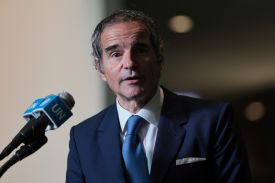 Grossi is seen from the mid-torso on upward as he speaks into a microphone. He wears a dark blue suit and a lighter blue tie, and a shadow cuts across his face, mostly obscuring the right half of it.