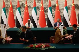 Xi and Sheikh Mohammed bin Zayed sit at a long table, with UAE and Chinese flags hanging in the background. Other officials move around in the foreground, shaking hands and holding folders of papers.