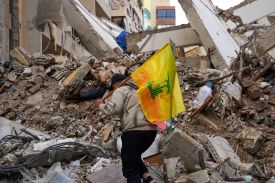 A man wearing a gray hoodie and carrying a yellow Hezbollah flag walks over the rubble of a damaged building.