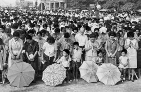 A large crowd of people bow their heads as they observe a minute of silence during the peace memorial ceremony on the 22nd anniversary of the Hiroshima attack at Hiroshima Peace Memorial Park on Aug. 6, 1967. Umbrellas and some small children are seen in the front row.