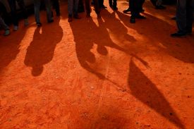 The feet of a small crowd of people are seen at the top of the image, shadows of the rest of their bodies cast on the reddish dirt ground below. One person's arms are raised as they wave a flag.