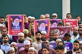 Dozens of men stand in a crowd outdoors with serious expressions on their faces. Some of them hold up computer-printed posters with messages such as "Why was Ayatollah Khamenei killed? America and Israel must answer." Another sign says "Muslim world unite!"