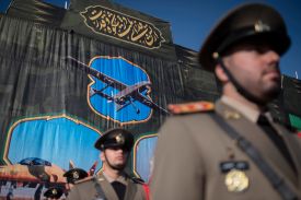Officers in uniform with formal hats stand under a banner with words in Farsi and the image of an unmanned drone.