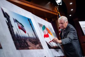 Graham, an elderly man with white hair wearing a business suit, draws with a marker on a photo of an Iranian flag flying outside somewhere.