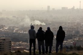 Silhouettes of four men are shown looking over a cityscape with smoke.