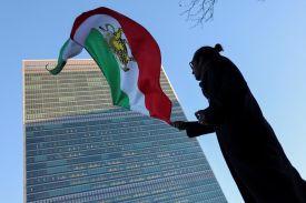A silhouetted person waves an older Iranian flag, which has the colors of green, white, and red, and the image of a yellow lion. In the background is the United Nations headquarters.