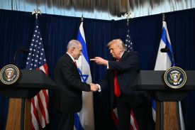 Trump and Netanyahu face each other and shake hands while standing near lecterns at the front of a press briefing room. Trump is pointing at Netanyahu's face, while Netanyahu smiles at him.