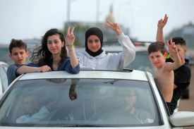 Five seemingly teenagers lean out of the open sunroof and windows of a car, several of them flashing peace signs with their hands. The oldest girl wears a hijab while another has her hair loose; one of the youngest boys is shirtless. Two adults are visible through the windshield in the front seat of the car.