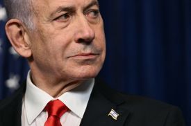 A close headshot of Netanyahu wearing a suit and tie and Israeli flag pin looks out of the side of his eyes.