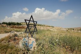 A Star of David made up of black-painted wooden planks stands in a field, surrounded by tall grasses and scattered rocks. Fluffy white clouds drift by against a blue sky overhead.