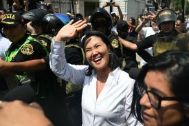 Flanked by police offers, Peruvian presidential candidate Keiko Fujimori waves to a crowd. She is wearing a white shirt and a crucifix necklace.