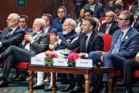 The four leaders sit in red upholstered chairs in the front row of an audience of other officials and aides. Modi and Macron smile and hold hands together, whereas Lulas has a serious expression and his hands clasped in front of him.