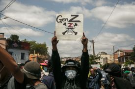 A person wearing a medical mask holds up a poster that reads "GenZ" with a skull and crossbones underneath. They are in a crowd of other people.
