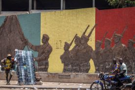Two people on a motorcycle and one person standing by a rack of sunglasses are shown before a mural depicting soldiers.
