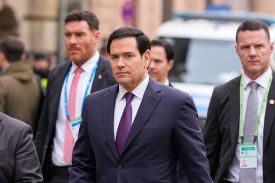 Rubio dressed in a blue suit and red and blue tie walks down a street surrounded by a group of men wearing conference badges.