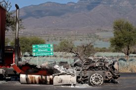 The debris of a burned truck is seen on a road in Mexico. Behind it are green highway signs and mountainous terrain beneath a clear blue sky.