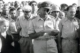 A black and white photograph captures a somber gathering of people outdoors. In the center, a man in a military uniform and a peaked cap looks down while holding and reading from a small piece of paper. He wears a dark patch over his left eye.