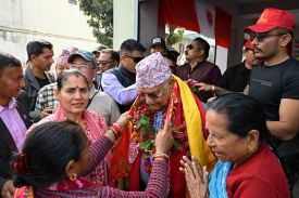 Oli, an older Nepalese politician, stands among a crowd of supporters as a few of them place a garland over his head.