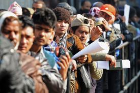 Dozens of people, mostly men, are seen leaning against a railing as they stand in line. Several of the men hold passports or bundles of paper.