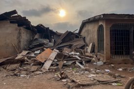 A pile of rubble from a destroyed building sits between two mostly intact single-story structures. The sun is visible overhead, low in the sky as it permeates through gray partial cloud cover.