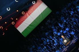 Orban is seen with the camera tilted at an angle as he speaks at a podium beneath a Hungarian flag. Several rows of a crowd of spectators are visible seated behind him.