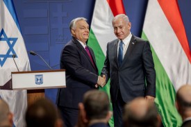 Netanyahu and Orban stand side by side as they shake hands and smile toward an out-of-focus seated audience in the foreground. Behind them are a podium with the Israeli state emblem as well as Israeli and Hungarian flags. Both men are wearing suits and ties.