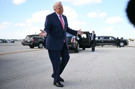 Trump gestures with both hands, palms lifted to the sky, as he walks across an airport tarmac beneath a light blue sky with a few clouds. Secret Service agents wait by black cars in the background.