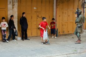 An armed Israeli soldier stands guard near children standing by closed shop doors.