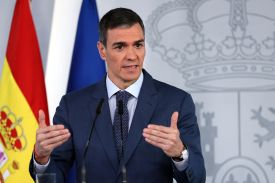 Flanked by the Spanish and European Union flags, Sanchez is seen from the chest upward as he speaks at a podium. He wears a dark suit and tie and gestures with both hands as he talks.