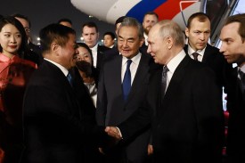 Chinese Foreign Minister Wang Yi shakes hands with Russian President Vladimir Putin while welcoming him to Beijing Capital International Airport. Several other officials stand in a crowd behind and around them.