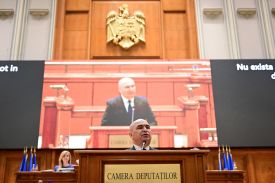 Romanian Prime Minister Ilie Bolojan stands in front of a podium to address parliament. A large screen airing his speech live displays behind him.