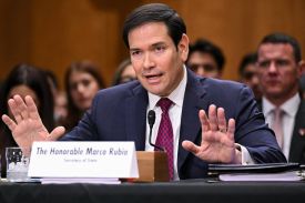 Secretary of State Marco Rubio holds up his hands from a seated position and from behind a name placard as he speaks with his back to attendees at a Senate Foreign Relations Committee hearing.