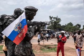 A Russian flags from a statue of a soldier holding a gun. People holding cell phones appear to take pictures at right as cars, motorbikes, and people are seen on a street behind them.