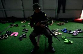 A guard with a gun sits in a chair in the shadows. Behind him are dozens of sandals.