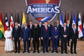 Trump stands with a dozen other leaders, all dressed in business formal attire. Behind them are the flags of several countries in the region as well as a large seal showing the name of the summit.