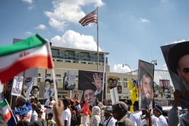 People are seen outside the U.S. consulate, with a U.S. flag flying above it. On the fence along the consulate are posters of Iranian leaders.